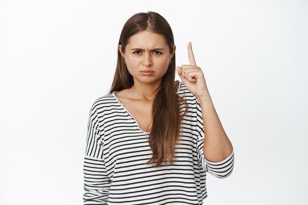 Разочарованная молодая женщина, хмурящаяся и указывающая пальцем на что-то плохое, стоящая в полосатой блузке на белом фоне