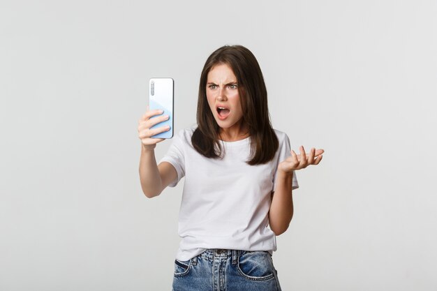 Frustrated young woman having argument on video call, holding smartphone, standing white.