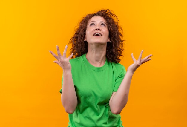 Frustrated young woman with short curly hair in green t-shirt looking up with raised hands 