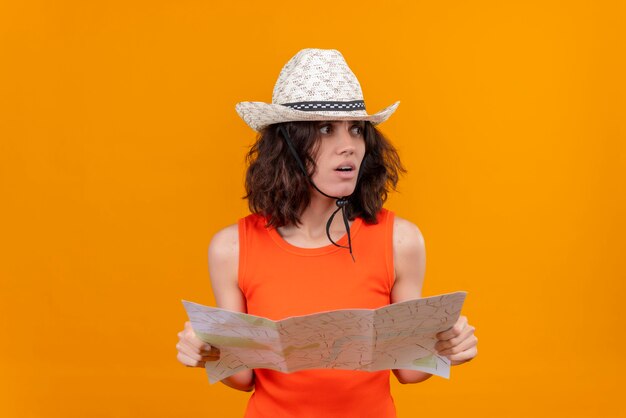 A frustrated young woman with short hair in an orange shirt wearing sun hat holding map and looking side 