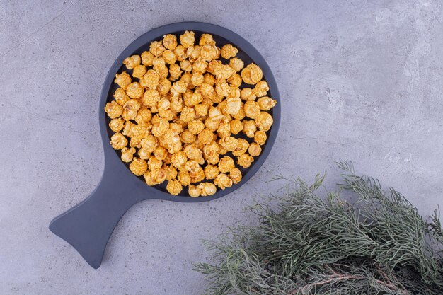Frying pan filled with caramel coated popcorn next to evergreen branches on marble background. High quality photo