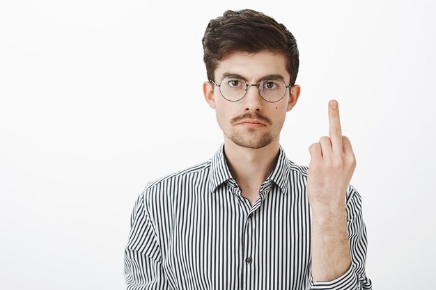 Fuck off, man, I am not your slave. Annoyed serious adult male with bristle and moustache, showing middle finger and staring with calm expression, being fed up of mocking over gray wall