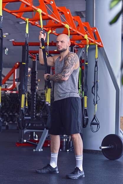 The full body image of athletic bearded tattooed, shaved head male doing trx straps exercises in a gym club.