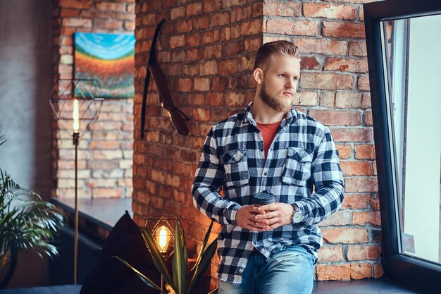 The full body image of blond hipster male drinks coffee in a room with loft interior.