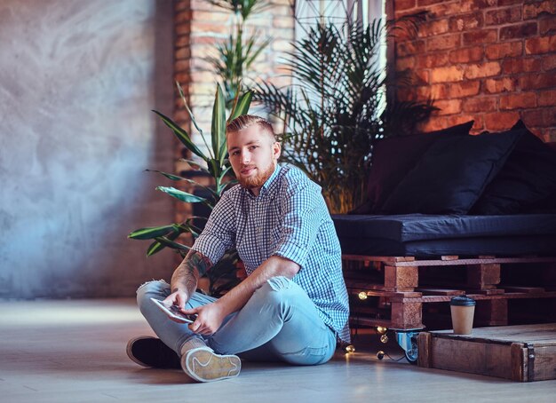 The full body image of blond stylish male dressed in a fleece shirt and jeans using a tablet PC in a living room.