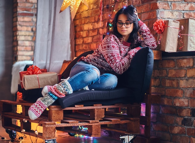 Full body image of brunette female in eyeglasses dressed in a jeans and a red sweater posing on a wooden sofa in a room with Christmas decoration.