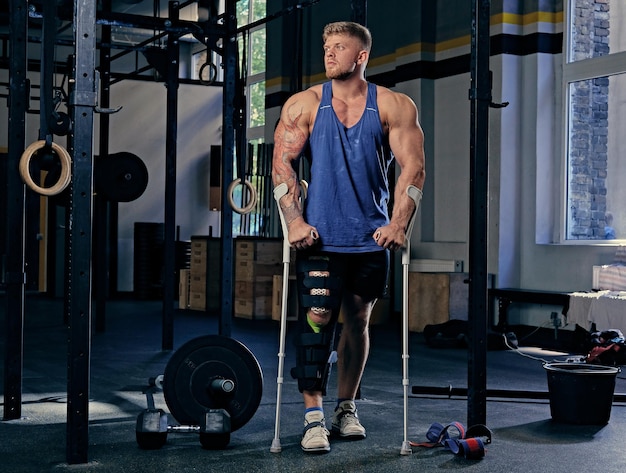 Full body image of huge bodybuilder on crutches in a gym club.