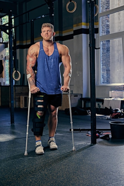 Full body image of huge bodybuilder on crutches in a gym club.