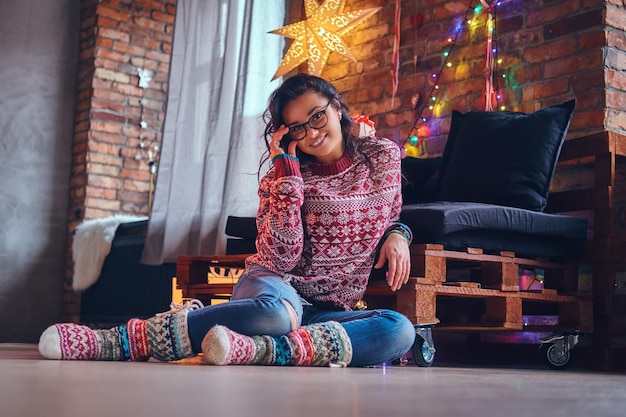 Full body image of sexy brunette femalesits on a floor in a room with Christmas decoration.