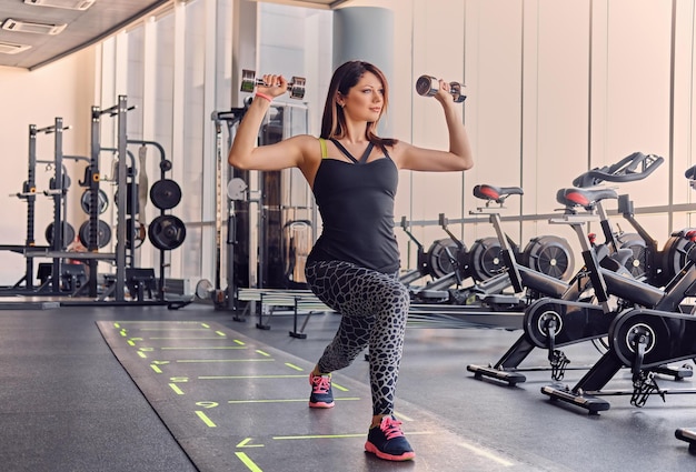 The full body image of sporty female holds dumbbells and doing squats in a gym.