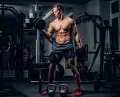 Free photo full body portrait of athletic shirtless male doing biceps workouts with dumbbells in a gym club.