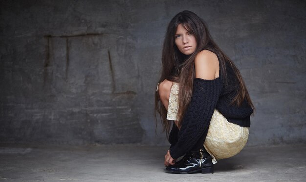 Full body portrait of brunette fashion woman who sitting on the floor over grey wall.