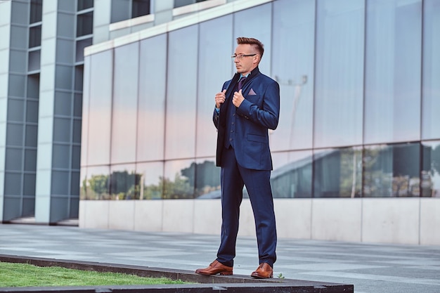 Full body portrait of a confident man in elegant suit corrects his jacket while standing against skyscraper background.