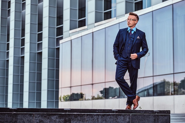 Full body portrait of a confident man in an elegant suit posing with hand in pocket against a skyscraper background.