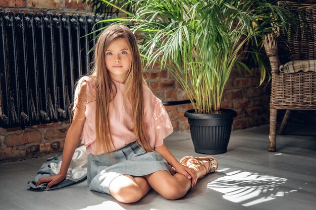 Full body portrait of a girl sits on a floor with plants on background.