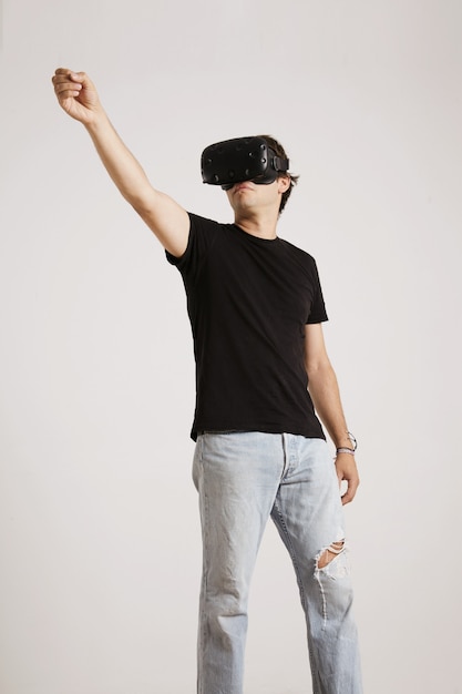 Full body portrait of a man in torn jeans and unlabeled black t-shirt wearing VR headset holding something high up isolated on white