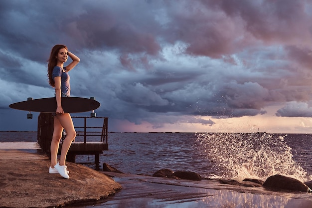 Полный портрет чувственной девушки, держащей скейтборд, стоящей на пляже и наслаждающейся удивительной темной облачной погодой во время заката.