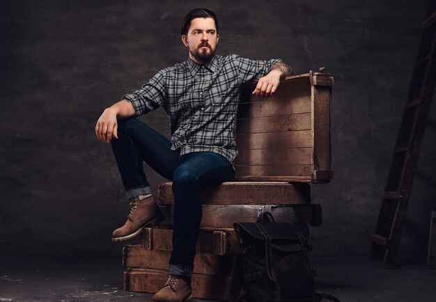 Full body portrait of a tattoed middle age hipster man dressed in a checkered shirt and jeans, sitting on wooden boxes in a studio. Isolated on a dark textured background.