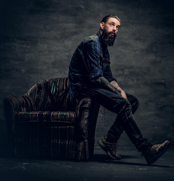 Full body portrait of tattooed, bearded male dressed in a plaid flannel shirt sits on a chair over dark grey background.