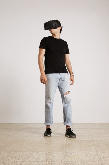 VRヘッドセット、黒いラベルのないTシャツ、白い壁と明るいフローリングの床で部屋を見回す青い破れたジーンズの若い男性モデルの全身の肖像画