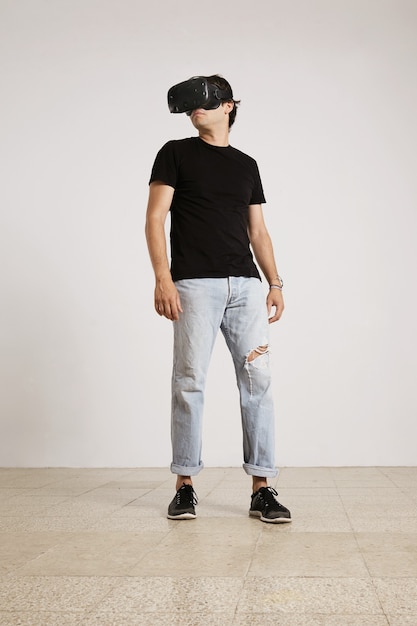 Full body portrait of a young male model in VR headset, black unlabeled t-shirt and blue torn jeans looking around the room with white walls and light wooden floor