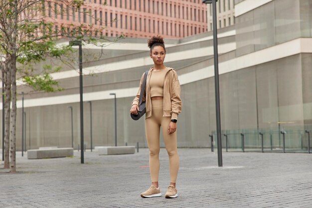 Full body shot of thoughtful sporty slim woman dressed in sportswear carries karemat looks away outdoors