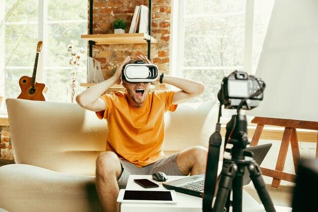 Full of emotions. Caucasian male blogger with professional camera recording video review of VR glasses at home. Blogging, videoblog, vlogging. Man using virtual reality headset while streaming live.