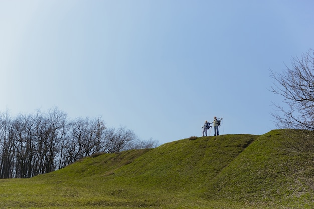 Full and free breathing. Aged family couple of man and woman in tourist outfit walking at green lawn near by trees in sunny day. Concept of tourism, healthy lifestyle, relaxation and togetherness.