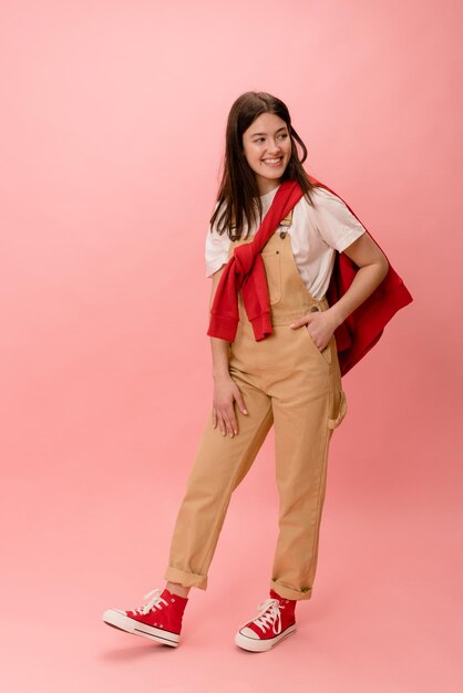 In full growth stylish young european brunette girl in overalls holds hand in pocket on pink background Lifestyle different emotions leisure concept