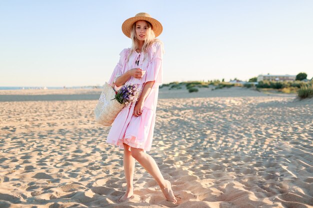 Изображение в полный рост белокурой девушки в милом розовом платье, танцующей и имеющей фу на пляже. Держит соломенный мешок и цветы.