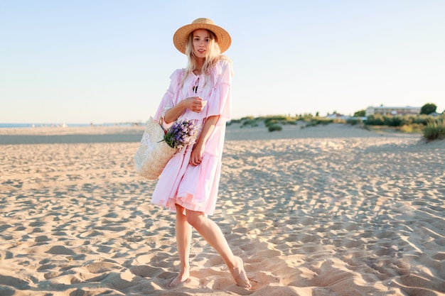 Full height  image of   blond   girl in cute pink  dress dancing and having fu on  the beach. Holding straw bag  and  flowers.