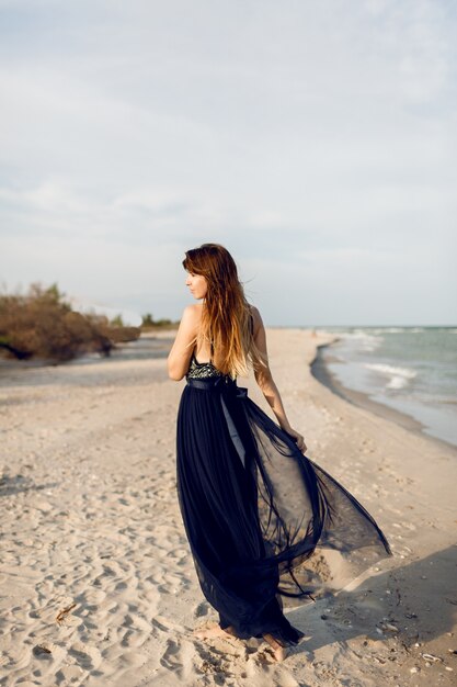 해변에서 포즈를 취하는 우아한 럭셔리 드레스에 유행 여자의 전체 높이 이미지. 뒤에서 봅니다. 긴 머리카락.