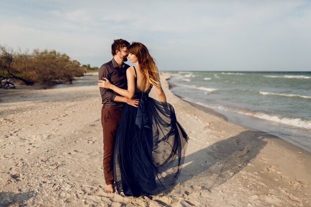 海の近くのイブニングビーチでロマンチックなカップルを受け入れる高さの完全なイメージ。優しさと彼女のボーイフレンドを抱き締める青いロングドレスの見事な女性。ハネムーン。