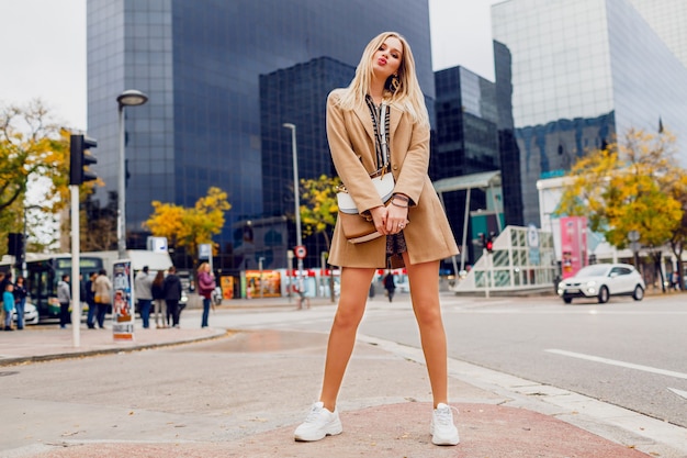 Full height portrait of pretty blond woman posing over urban street. Wearing beige coat and white sneakers. Trendy accessories. Carefree lady walking along the street.