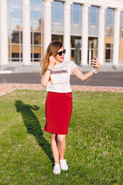 Full-height portrait of a standing smiling young girl holding a smartphone and making a selfie