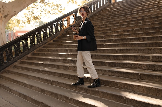 Полноценная женщина в ботинках, куртке и белых брюках держит телефон и чашку с тестом снаружи. Современная женщина в очках позирует