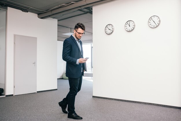 Full-lengh photo of attractive guy in glasses walking in office. He wears blue shirt, dark jacket, jeans, black shoes and beard. He is typing on phone.