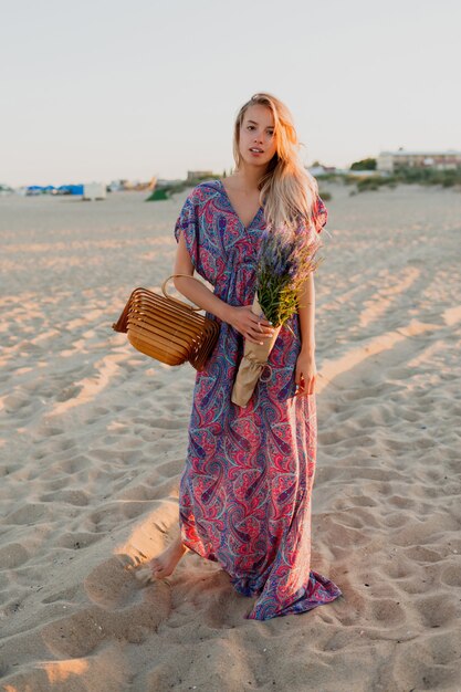 ビーチを歩いているラベンダーの花束を持つかなり金髪の女性の完全な長さの画像。夕焼けの色。