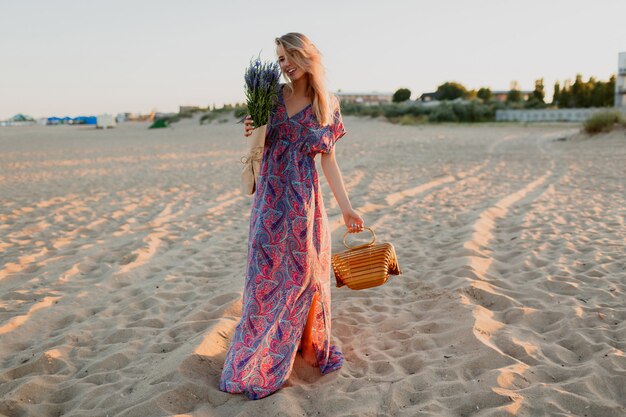 ビーチを歩いているラベンダーの花束を持つかなり金髪の女性の完全な長さの画像。夕焼けの色。