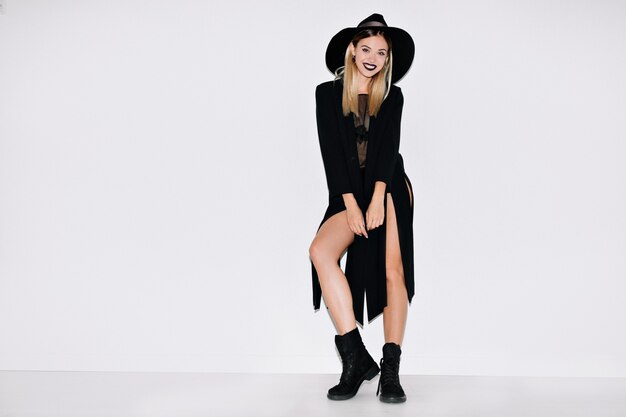 Full-lenght portrait of cheerful stylish  modern woman wearing long black jaket and hat, boots standing over isolated wall