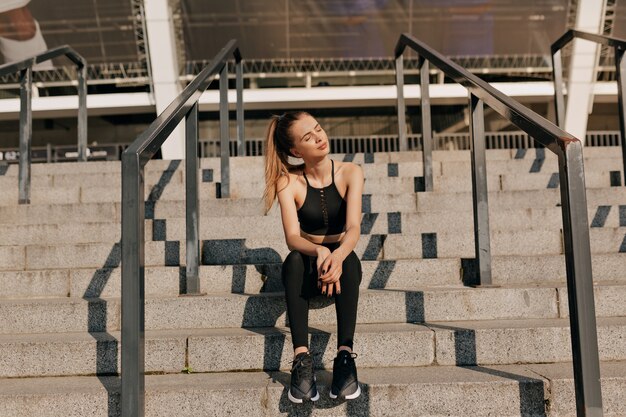 Full-lenght portrait of young fit woman with collected hair sitting outside