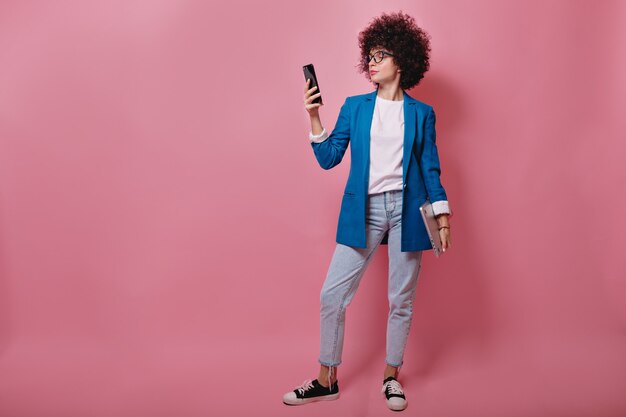 Full-lenght portrait of young successful woman with short afro hairstyle in blue jacket and jeans using smartphone over pink wall