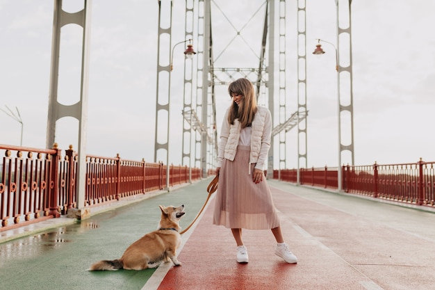 Full-lenght shot of stylish european woman wearing pink skirt and white jacket walking with a corgi dog in morning sunny city.