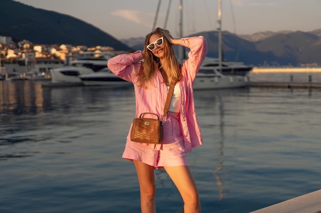 Полноразмерный солнечный портрет стильной женщины, гуляющей в роскошном яхт-клубе в Черногории, наслаждающейся отдыхом на закате