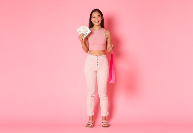 夏の服で魅力的な笑顔のアジアの女の子の完全な長さ、お金と買い物袋を保持している、高価なものを購入する、モールを歩いて楽しんでいる、ピンクの壁に立っている