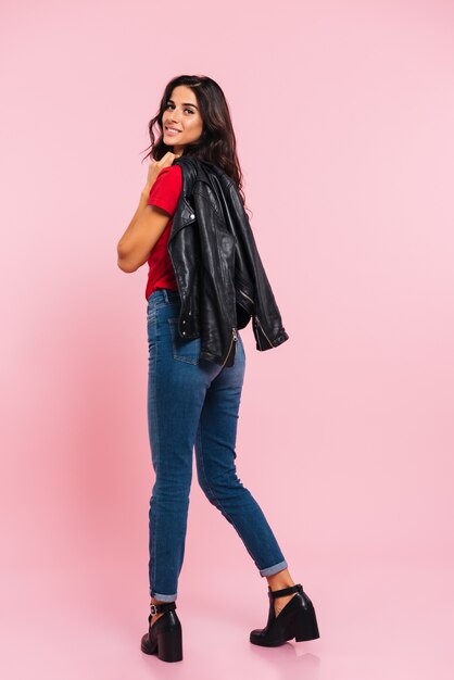 Full length back view of smiling brunette posing in studio and looking at the camera over pink 