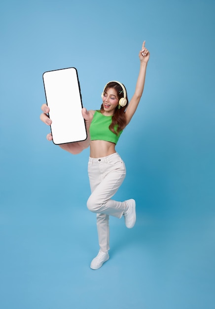 Full length beautiful asian teen woman showing smartphone mockup of blank screen and listening music