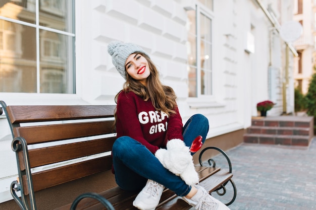 니트 모자, 청바지와 거리에 벤치에 앉아 흰 장갑에 긴 머리를 가진 전체 길이 아름 다운 어린 소녀. 그녀는 카라멜 하트를 들고 웃고 있습니다.