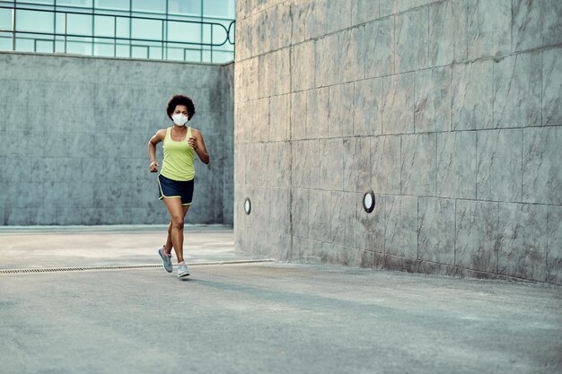 Full length of black sportswoman with face mask running in the city