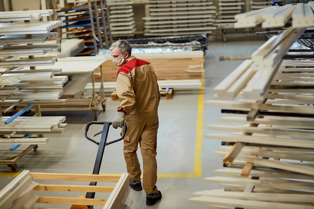 Full length of carpenter with face mask pulling pallet jack in a workshop